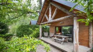 Blockhütte mit überdachter Veranda und Picknicktisch, umgeben von Bäumen im Huttopia Rambouillet, Frankreich.