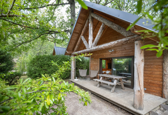 Blockhütte mit überdachter Veranda und Picknicktisch, umgeben von Bäumen im Huttopia Rambouillet, Frankreich.