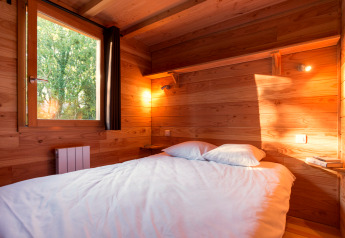 Cozy bedroom in a cabin with a wooden interior, double bed, shelf, and sunlight coming through the window.