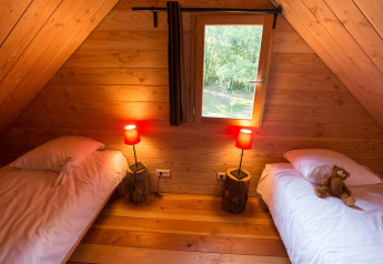 Cozy wooden cabin bedroom with two single beds, plush toys on each bed, warm lamps, and a window view.
