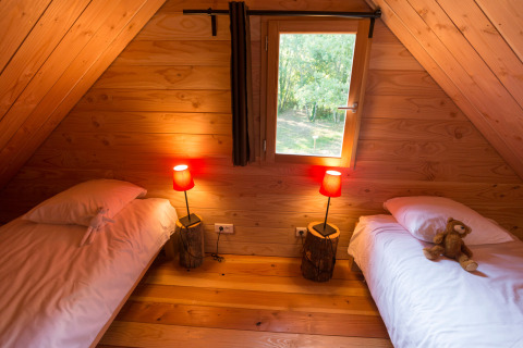 Cozy wooden cabin bedroom with two single beds, plush toys on each bed, warm lamps, and a window view.