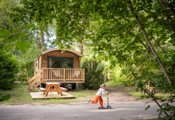 A child rides a scooter in front of a cozy wooden cabin called Gypsy caravan surrounded by lush forest.