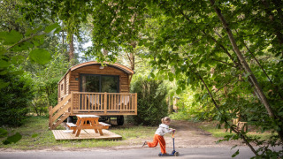 Un niño monta un patinete frente a una acogedora cabaña de madera llamada Gypsy caravan en el bosque.