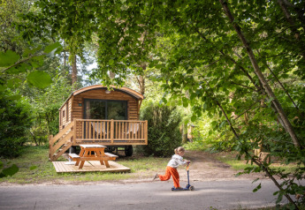 Een kind rijdt op een step voor een houten huisje genaamd Gypsy caravan in een groene bosrijke omgeving.
