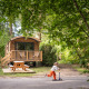 Et barn på løbehjul foran en hyggelig træhytte kaldet Gypsy caravan midt i en grøn skov.