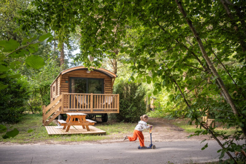 Un niño monta un patinete frente a una acogedora cabaña de madera llamada Gypsy caravan en el bosque.