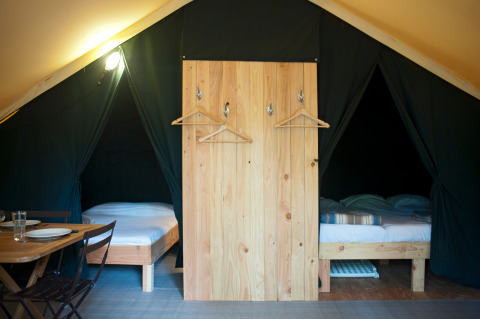 Interior de tienda safari con dos camas individuales, biombo de madera con perchas y mesa de comedor.