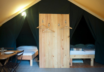 Interior de tienda safari con dos camas individuales, biombo de madera con perchas y mesa de comedor.