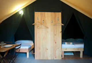 Intérieur d’une tente safari avec deux lits simples, séparateur en bois, patères et table à manger.