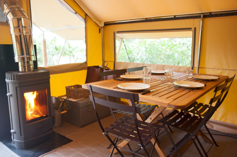 Vista interior de la tienda safari Tente Trappeur con mesa de comedor y estufa de leña en Huttopia Rambouillet, Francia.