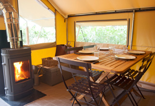Vista interior de la tienda safari Tente Trappeur con mesa de comedor y estufa de leña en Huttopia Rambouillet, Francia.