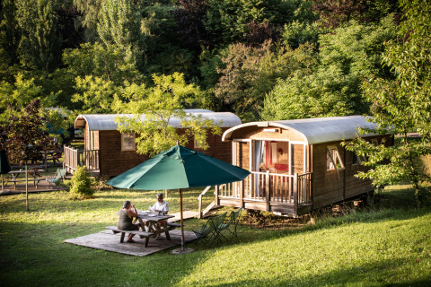 Two people sit at a table under an umbrella in front of wooden cabins at Huttopia Sarlat in France.