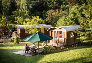 Two people sit at a table under an umbrella in front of wooden cabins at Huttopia Sarlat in France.