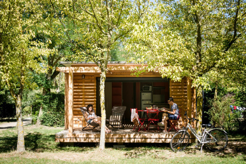 Deux personnes se détendent devant un chalet en bois entouré d’arbres à Huttopia Sarlat, Nouvelle-Aquitaine, France.