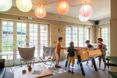 Niños juegan futbolín en una sala luminosa con farolillos de papel en Huttopia Sarlat, Nouvelle-Aquitaine, Francia.