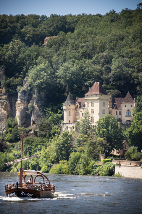 Un barco turístico navega por un río frente a un castillo rodeado de bosque en Huttopia Sarlat, Francia.