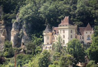 A tour boat sails on a river in front of a castle surrounded by lush forest at Huttopia Sarlat, France.