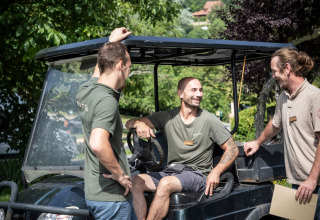 Tre mænd i afslappet samtale ved en golfvogn på Huttopia Sarlat feriested i Nouvelle-Aquitaine, Frankrig.