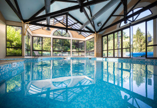 Indendørs swimmingpool med store vinduer og udsigt til grønne træer på Huttopia Sarlat i Frankrig.