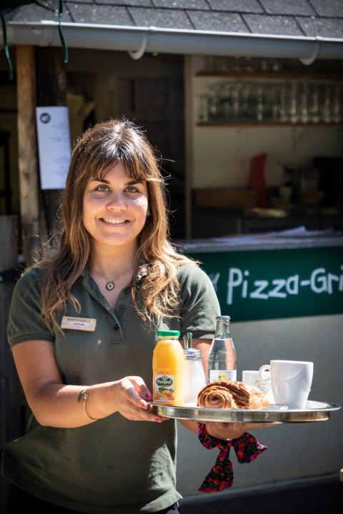 Mujer sirviendo desayuno con jugo, café y bollería en Huttopia Sarlat, Nouvelle-Aquitaine, Francia.
