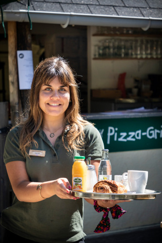 Femme servant un petit-déjeuner avec jus, café et viennoiserie à Huttopia Sarlat, Nouvelle-Aquitaine.