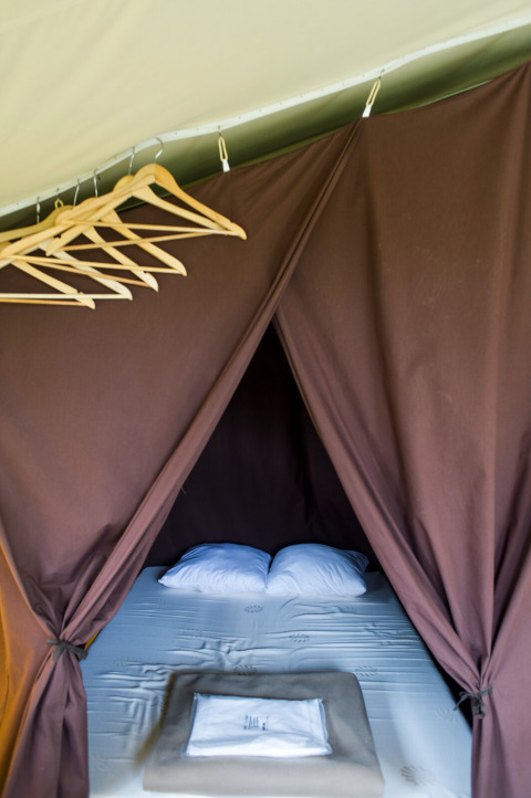 Interior de una tienda safari con cama doble, perchas y cortinas marrones en Huttopia Calvados - Normandie.