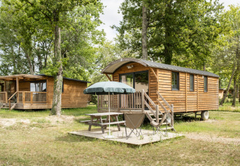 Cabañas de madera y una caravana gitana en Huttopia Rambouillet, Francia, con mesa y sombrilla en el exterior.