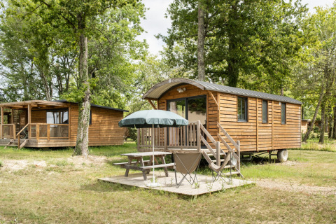 Cabañas de madera y una caravana gitana en Huttopia Rambouillet, Francia, con mesa y sombrilla en el exterior.