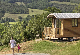 Holzige Zigeunerwagenhütte bei Huttopia Rambouillet in Frankreich mit grüner Hügellandschaft und Spaziergängern.
