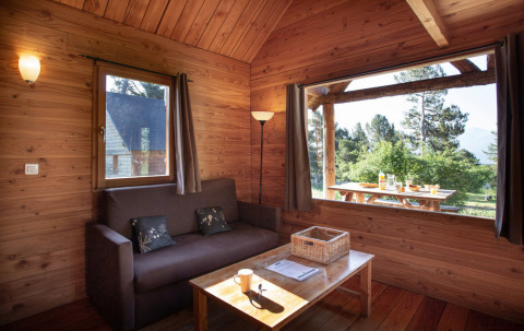 Intérieur d'une cabane en bois chaleureuse à Huttopia Rambouillet avec grande fenêtre sur la nature.