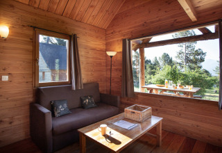 Innenansicht einer gemütlichen Holzhütte mit Sofa, Tisch und großem Fenster mit Blick ins Grüne.