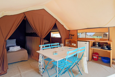 Interior of a safari tent featuring a dining area with blue chairs, beds, a kitchenette, and a small fridge.