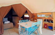 Interior of a safari tent featuring a dining area with blue chairs, beds, a kitchenette, and a small fridge.