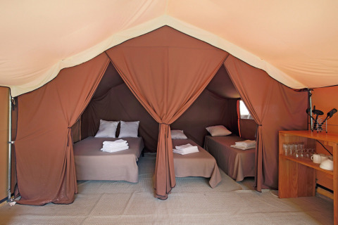 Interieur van een safaritent met drie opgemaakte bedden, kussens, handdoeken en een plank met serviesgoed.