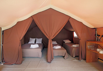 Interno di una tenda safari con tre letti pronti, cuscini, asciugamani piegati e una mensola con stoviglie.