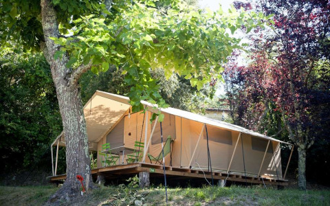Toile&bois tent Classic safari tent in a wooded area, with green chairs on a wooden deck and natural shade.