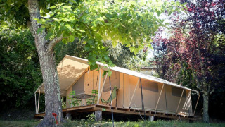 Safari-Zelt Toile&bois tent Classic im Grünen, mit Stühlen auf der Holzterrasse und Bäumen ringsum.