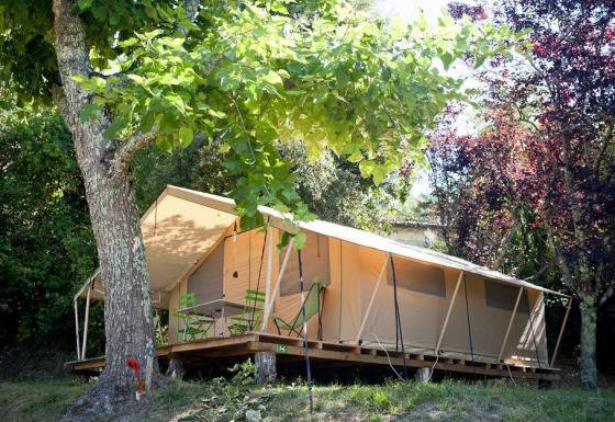 Tente safari Toile&bois tent Classic dans un cadre boisé, terrasse en bois et chaises vertes sous les arbres.