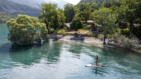 Persona practicando paddle surf en un lago junto a árboles y cabañas en Huttopia Lac de Serre Ponçon, Francia.
