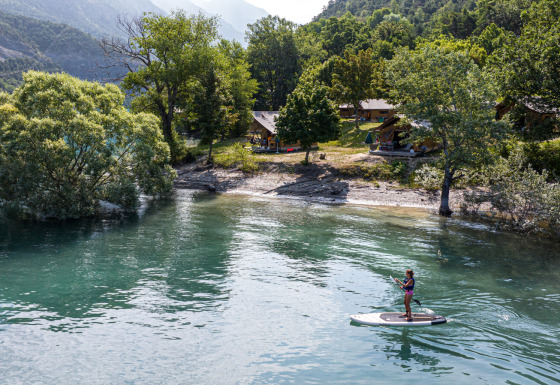 Persona practicando paddle surf en un lago junto a árboles y cabañas en Huttopia Lac de Serre Ponçon, Francia.