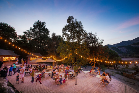 Reunión nocturna en Huttopia Lac de Serre Ponçon con luces de guirnalda y familias cenando al aire libre.