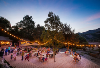 Avondtafereel bij Huttopia Lac de Serre Ponçon met feestverlichting en families op een houten terras in Frankrijk.