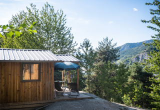 Houten chalet met terras en blauwe parasol bij Huttopia Lac de Serre Ponçon, met zicht op de groene bergen.
