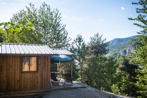 Cabaña de madera con terraza y sombrilla azul en Huttopia Lac de Serre Ponçon, rodeada de montañas verdes.