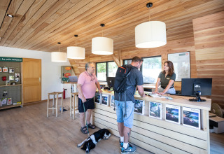 Receptionen i Huttopia Lac de Serre Ponçon feriepark, to gæster taler med personalet, hund på gulvet.