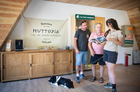 Receptioniste helpt twee mannen met hond bij Huttopia Lac de Serre Ponçon, Provence-Alpes-Côte d’Azur, Frankrijk.