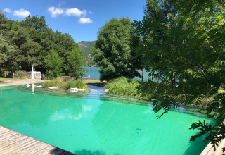 Natural swimming pool surrounded by lush trees, with lake view at Huttopia Lac de Serre Ponçon holiday park, France.