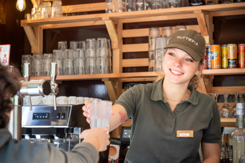 Smilende kvindelig bartender serverer en drink til gæst på Huttopia Lac de Serre Ponçon, feriepark i Frankrig.