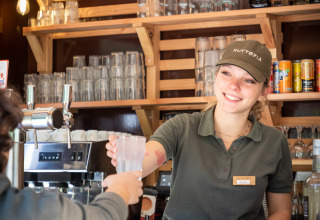 Smilende kvindelig bartender serverer en drink til gæst på Huttopia Lac de Serre Ponçon, feriepark i Frankrig.