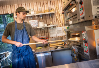 Un homme en tablier prépare une pizza dans la cuisine du Huttopia Lac de Serre Ponçon, en France.